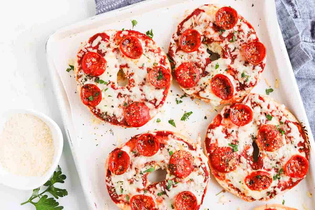 Four homemade pepperoni pizza bagels with melted cheese and pepperoni slices on a white tray, baked to perfection and garnished with chopped herbs.