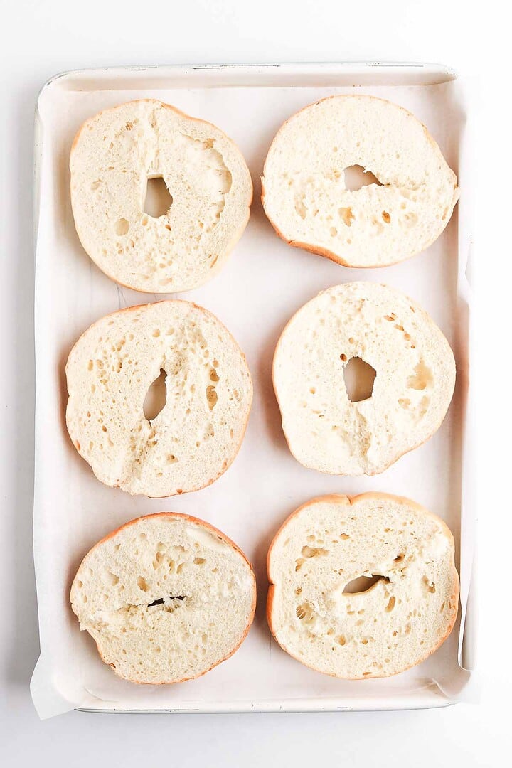 Six plain homemade bagel halves on a parchment-lined baking tray, arranged in two rows of three&mdash;perfect for turning into delicious Pepperoni Pizza Bagels.