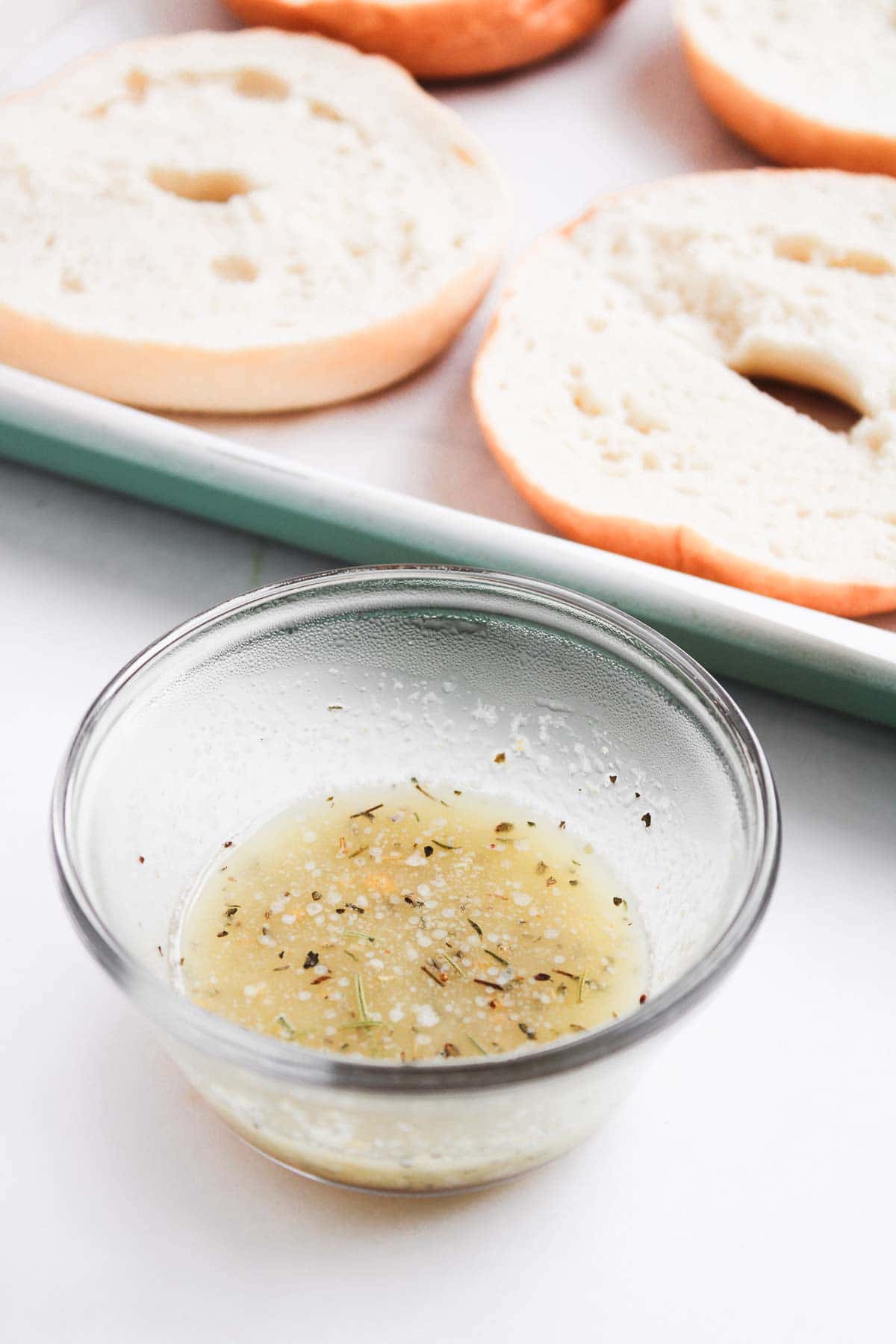 A glass bowl of homemade garlic herb butter sits in front of sliced plain bagels on a baking sheet, ready to be transformed into delicious baked Pepperoni Pizza Bagels.