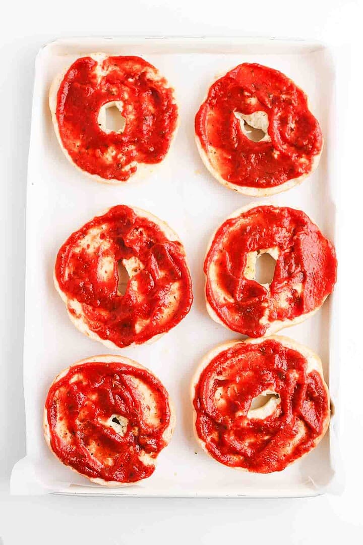 Six homemade pizza bagel halves with tomato sauce spread on top, arranged on a white baking sheet.
