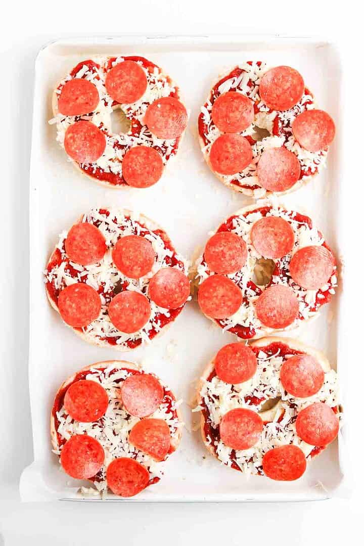 Six homemade pepperoni pizza bagels topped with tomato sauce, shredded cheese, and pepperoni slices are baked to perfection on a white baking tray.