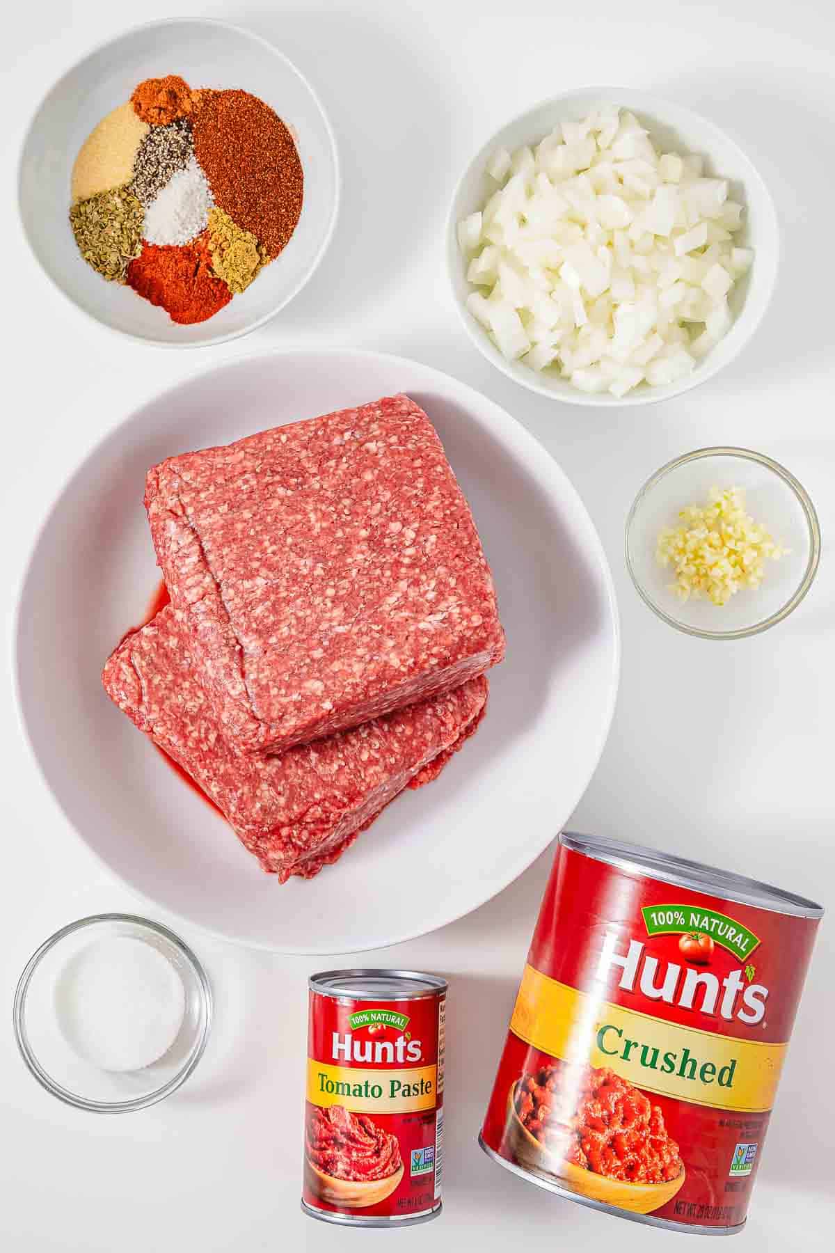 Raw ground beef, diced onions, garlic, spices, sugar, tomato paste, and crushed tomatoes arranged on a white surface&mdash;perfect ingredients for an easy Hot Dog Chili recipe.