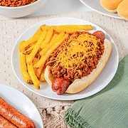 A chili cheese hot dog with fries on a plate, surrounded by extra chili, buns, and sausages—perfect for showcasing your favorite Hot Dog Chili as the ultimate Hot Dog Topping.