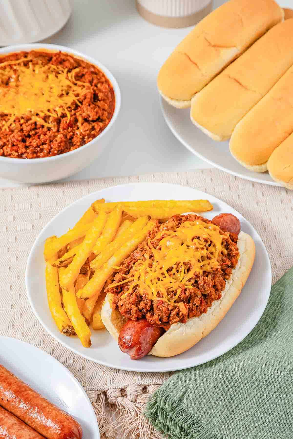 A chili cheese hot dog with fries on a plate, surrounded by extra chili, buns, and sausages&mdash;perfect for showcasing your favorite Hot Dog Chili as the ultimate Hot Dog Topping.