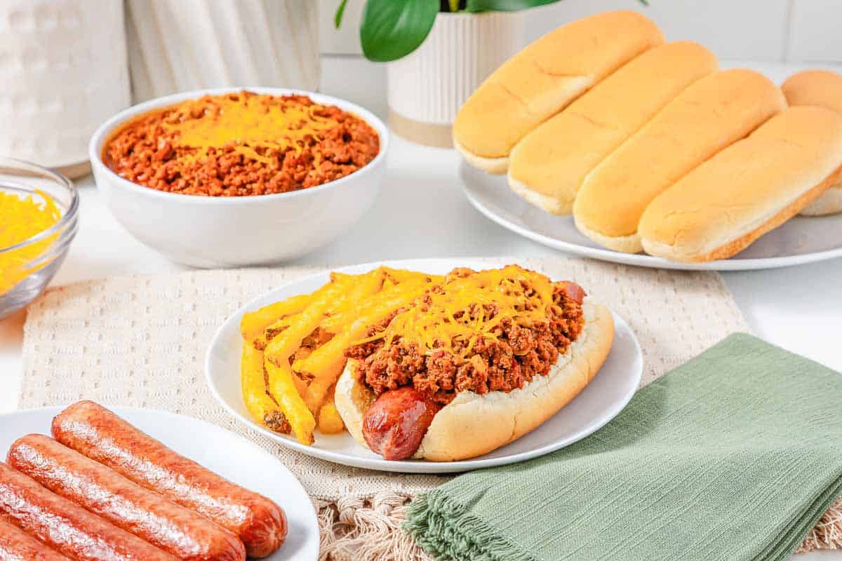 A chili dog with fries, hot dog buns, hot dogs, and bowls of Hot Dog Chili and cheese on a table&mdash;a classic spread featuring the perfect Hot Dog Topping.