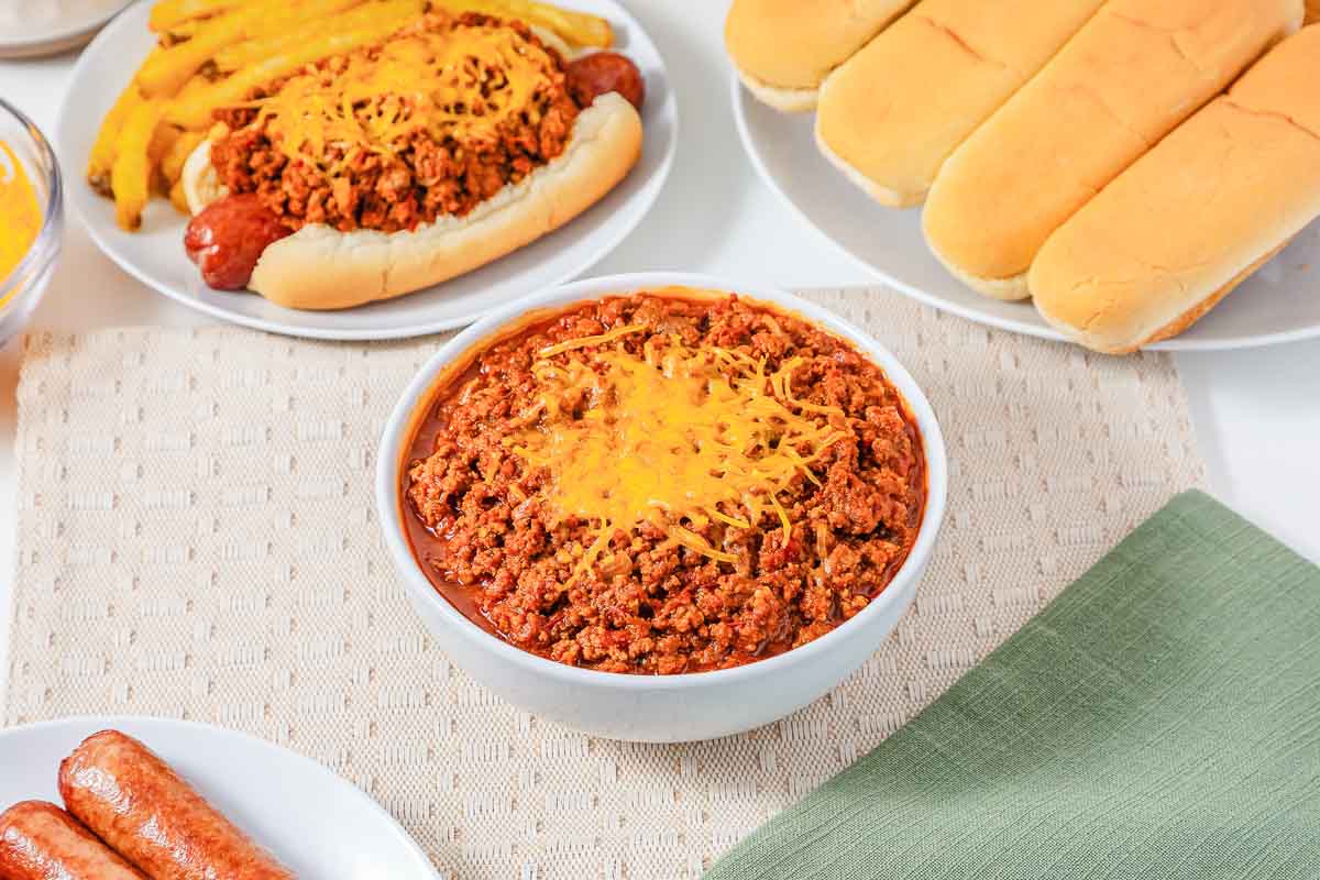 Bowl of hot dog chili topped with shredded cheese, with hot dogs, buns, and sausages on plates nearby.