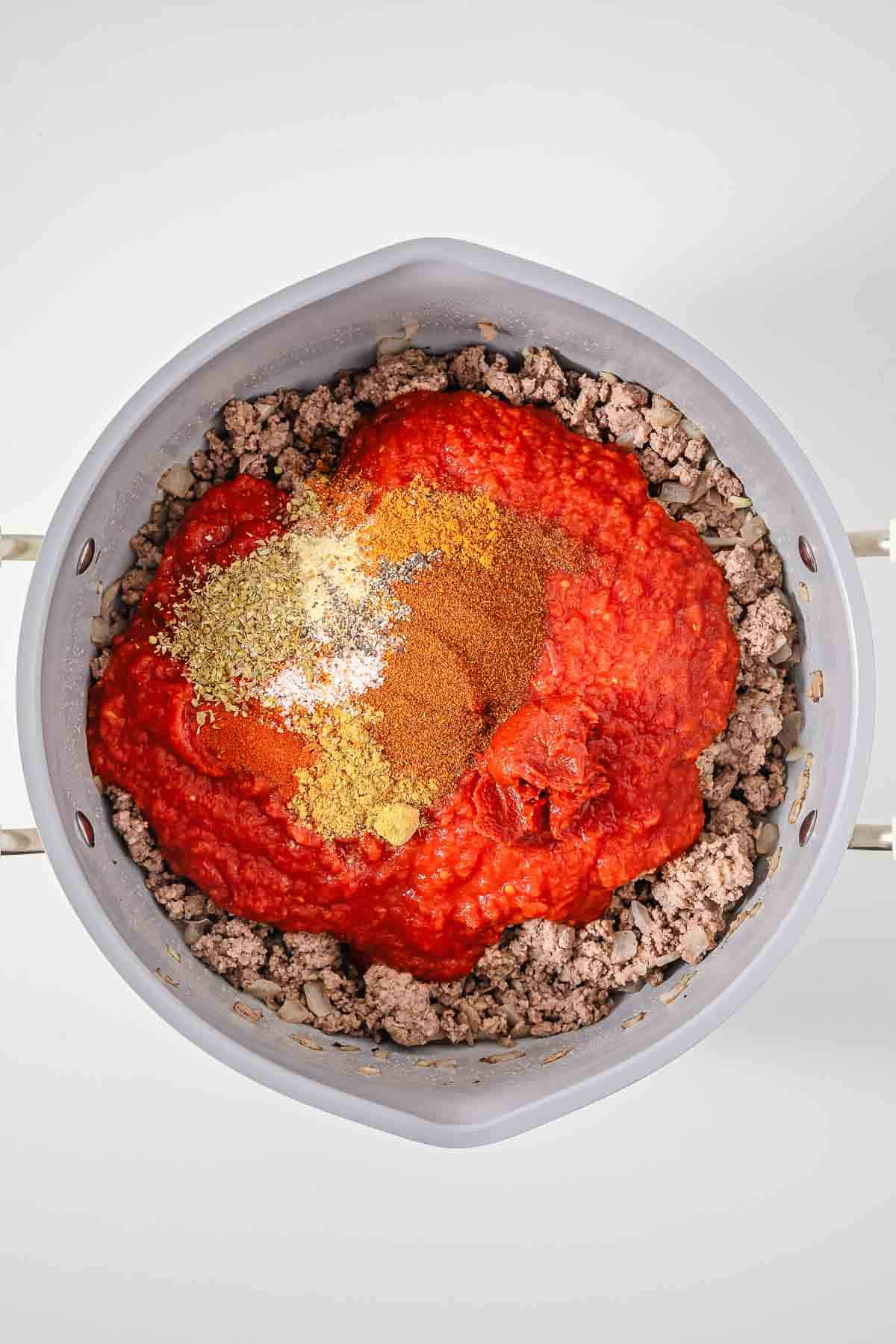 Ground meat in a pot topped with tomato sauce and various spices, perfect for an easy chili recipe or as a flavorful hot dog topping, ready to be mixed and cooked.