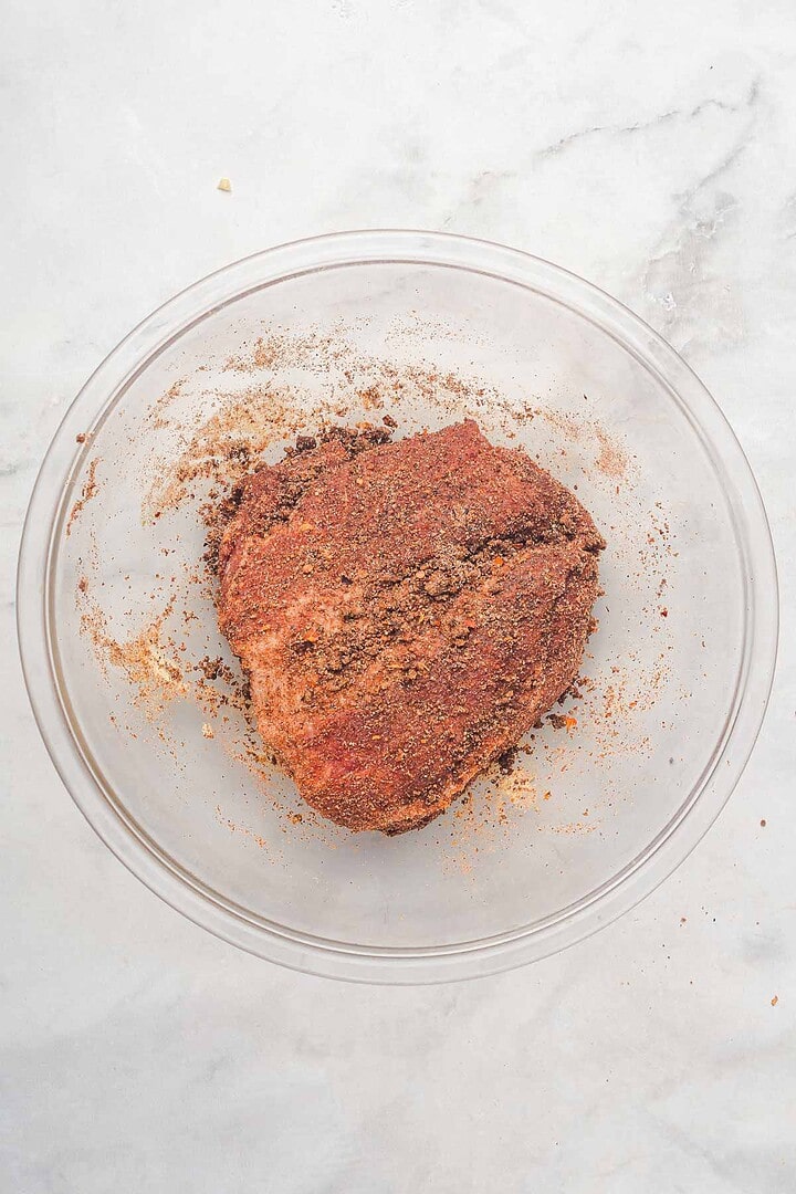 A glass bowl with a seasoned raw pork roast coated in dry rub, ready to become flavorful slow cooker Tex Mex pulled pork, sits on a light marble surface.