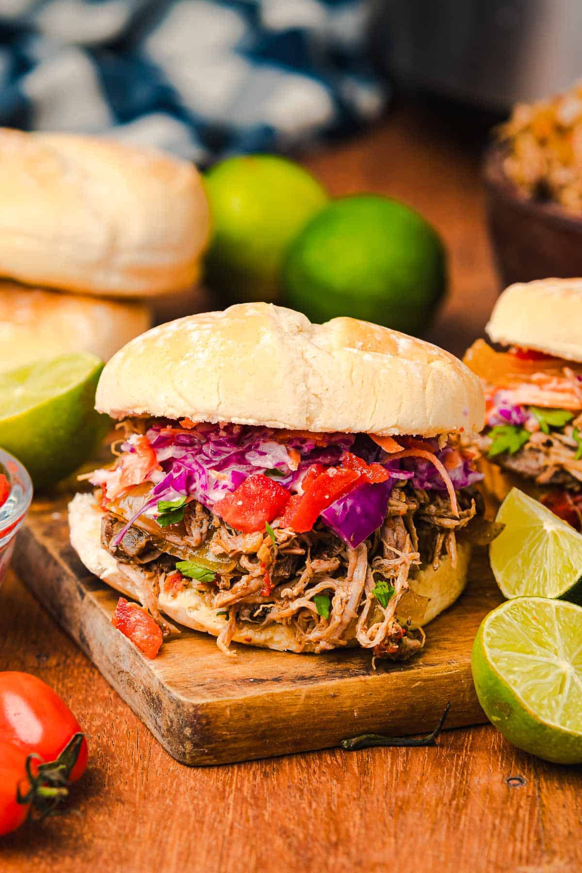 Slow cooker Tex Mex pulled pork sandwich with slaw and tomatoes on a bun, served with lime wedges on a wooden board.
