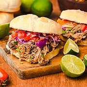 Slow cooker Tex Mex pulled pork sandwiches topped with coleslaw and tomatoes, served on a wooden board surrounded by limes and fresh veggies.