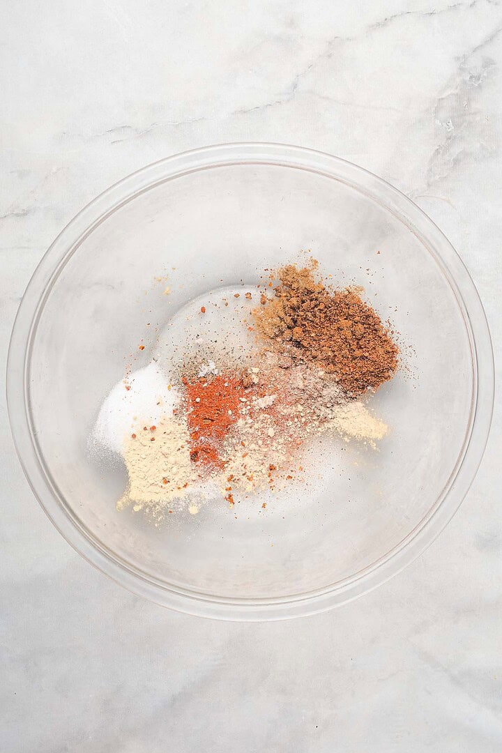 A glass bowl with various ground spices and seasonings on a light marble surface, perfect for preparing slow cooker Tex Mex pulled pork.