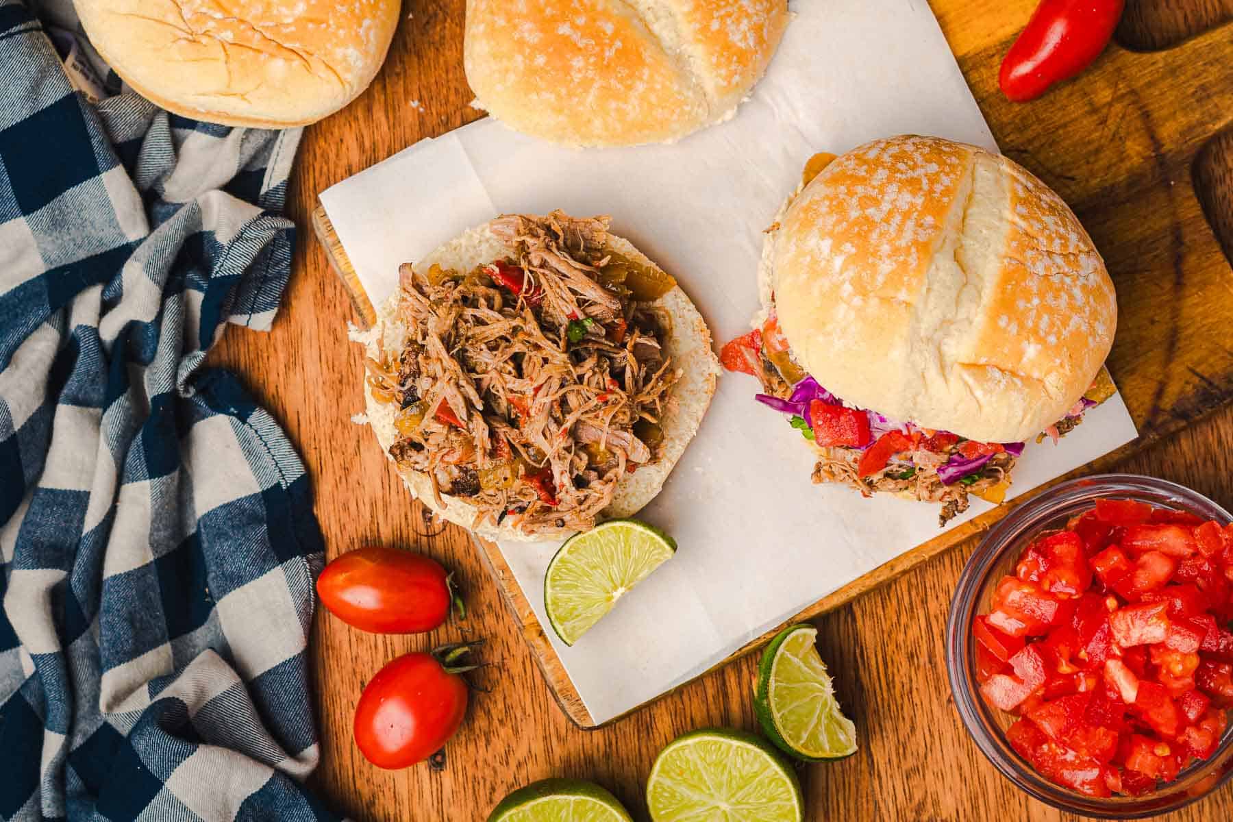Slow cooker Tex Mex pulled pork sandwiches with lime wedges, tomatoes, and a checkered napkin on a wooden table.
