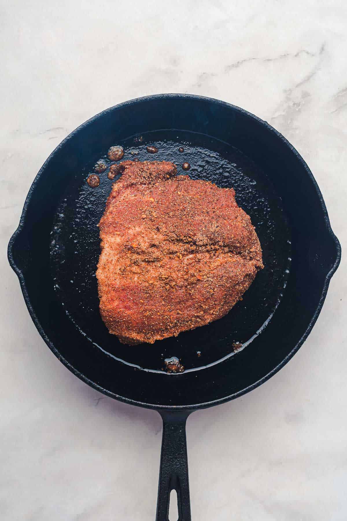 A seasoned piece of meat, reminiscent of slow cooker Tex Mex pulled pork, cooking in a black cast iron skillet on a light marble surface.