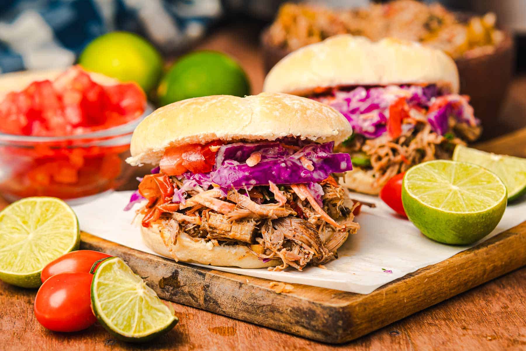 Two slow cooker Tex Mex pulled pork sandwiches with coleslaw and tomatoes on a board, served with fresh limes and tomatoes on the side.