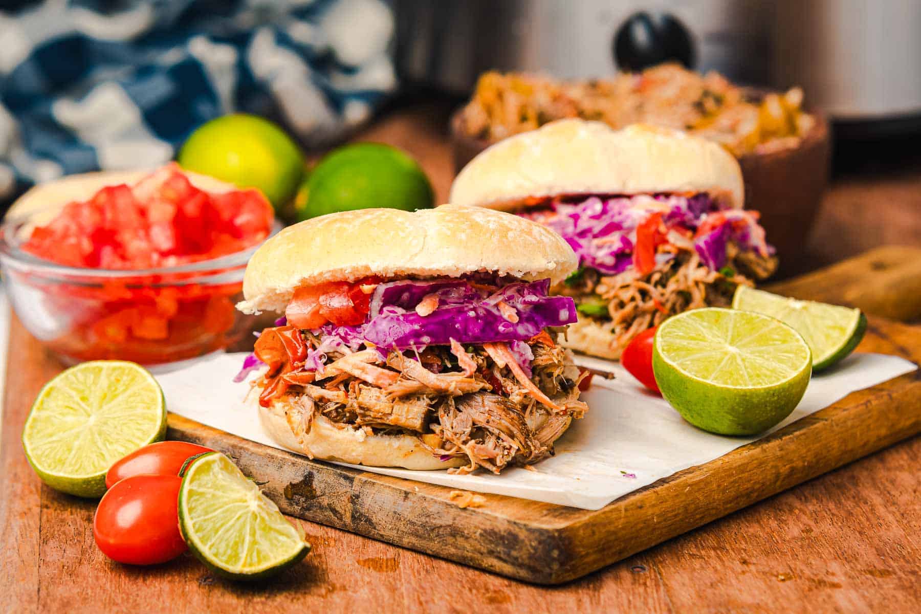 Two slow cooker Tex Mex pulled pork sandwiches with slaw on a board, garnished with lime halves and cherry tomatoes.