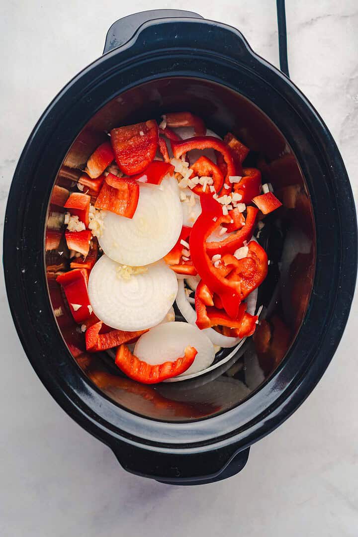 Sliced onions, red bell peppers, and minced garlic in a black slow cooker&mdash;perfect for starting your slow cooker Tex Mex pulled pork&mdash;on a marble surface.