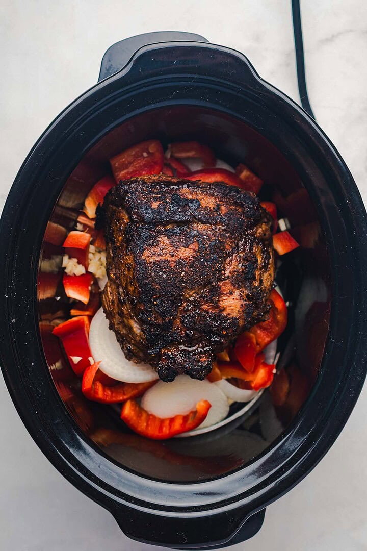 A seasoned slow cooker Tex Mex pulled pork roast rests on a bed of sliced onions and red bell peppers in a black slow cooker.