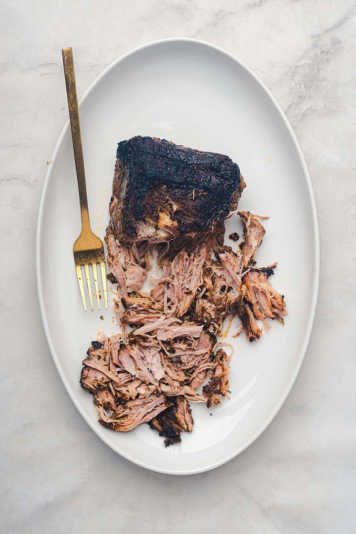 Slow cooker Tex Mex pulled pork, shredded and ready to serve with a golden fork on a white oval plate, set on a light-colored surface.