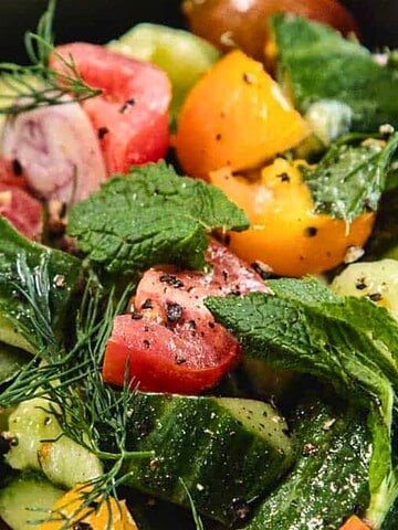 A fresh salad with tomatoes, cucumbers, mint, dill, and black pepper in a black bowl.
