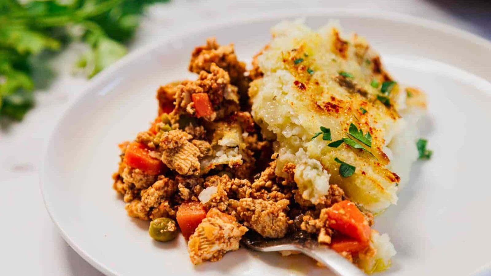 A serving of shepherd's pie with ground meat, peas, carrots, and mashed potatoes on a white plate.