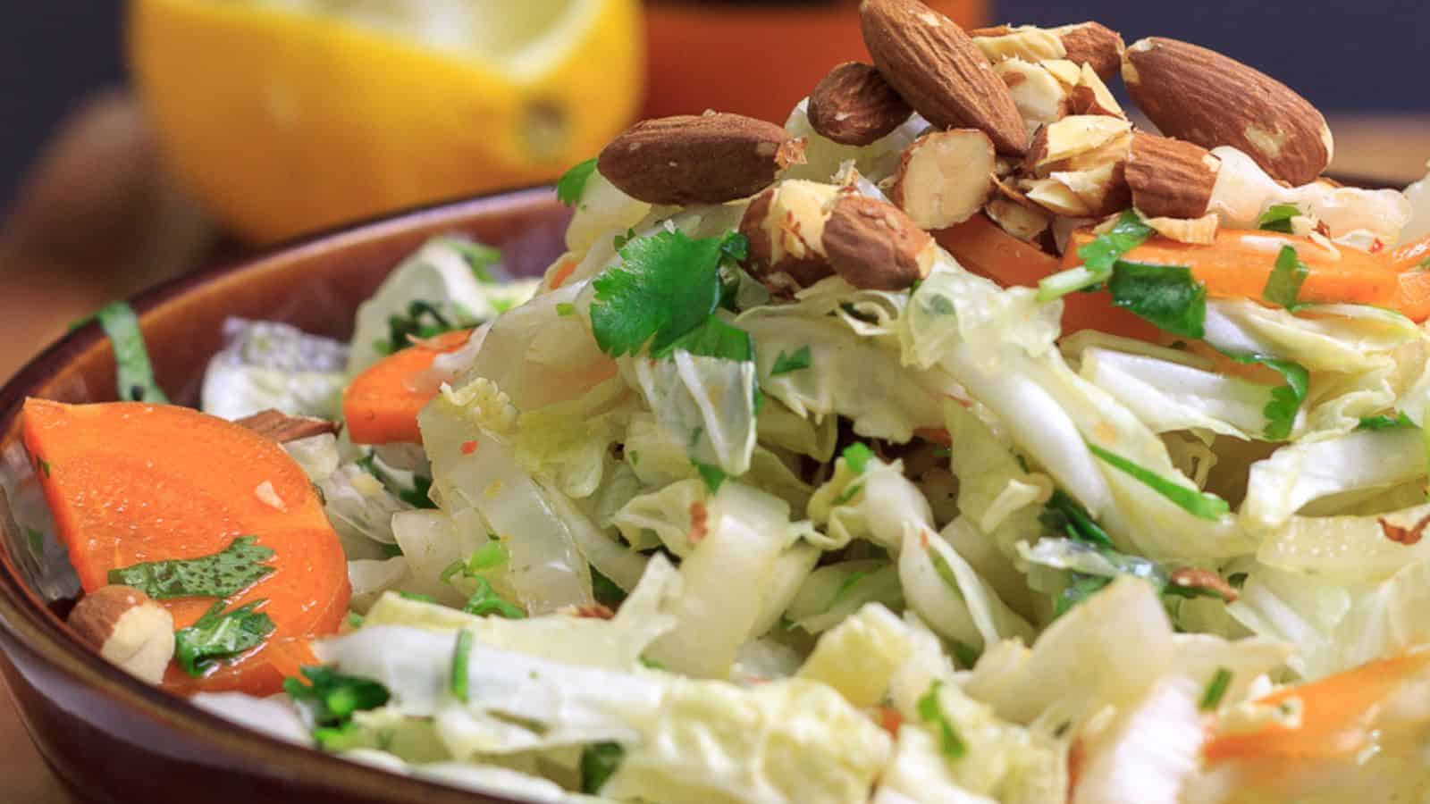 A bowl of cabbage salad with sliced carrots, chopped cilantro, and whole almonds, with a lemon in the background.