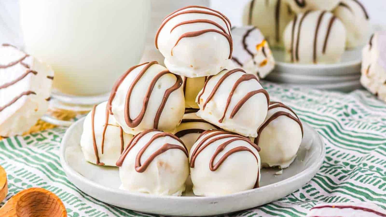 A plate of white chocolate truffles drizzled with chocolate, set on a green patterned cloth with wooden spoons nearby.