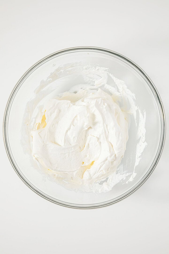 A glass bowl filled with fluffy white whipped cream on a white background, perfect for creating a classic Ambrosia or Fluff Salad recipe.