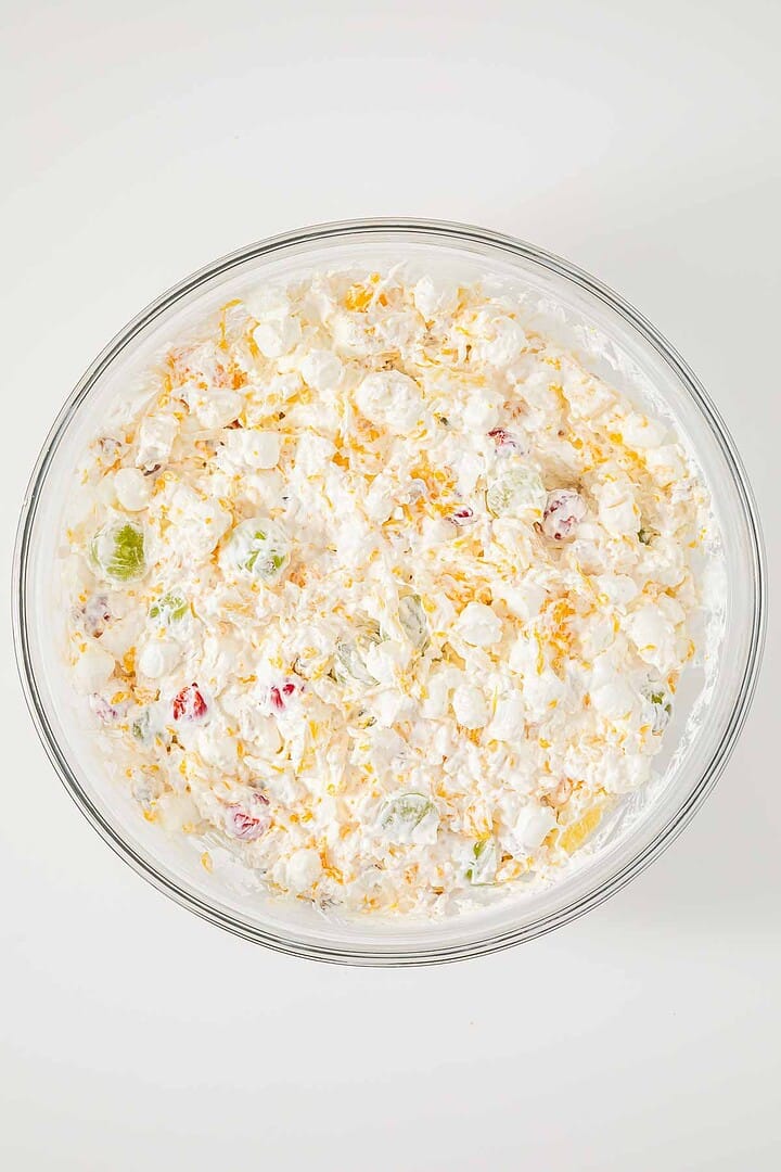 A glass bowl filled with creamy Ambrosia fruit salad, topped with shredded cheese, sits on a white surface.