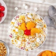 A bowl of ambrosia salad topped with fruit, cherries, and marshmallows sits surrounded by ingredients on a table, showcasing this classic fluff salad recipe.