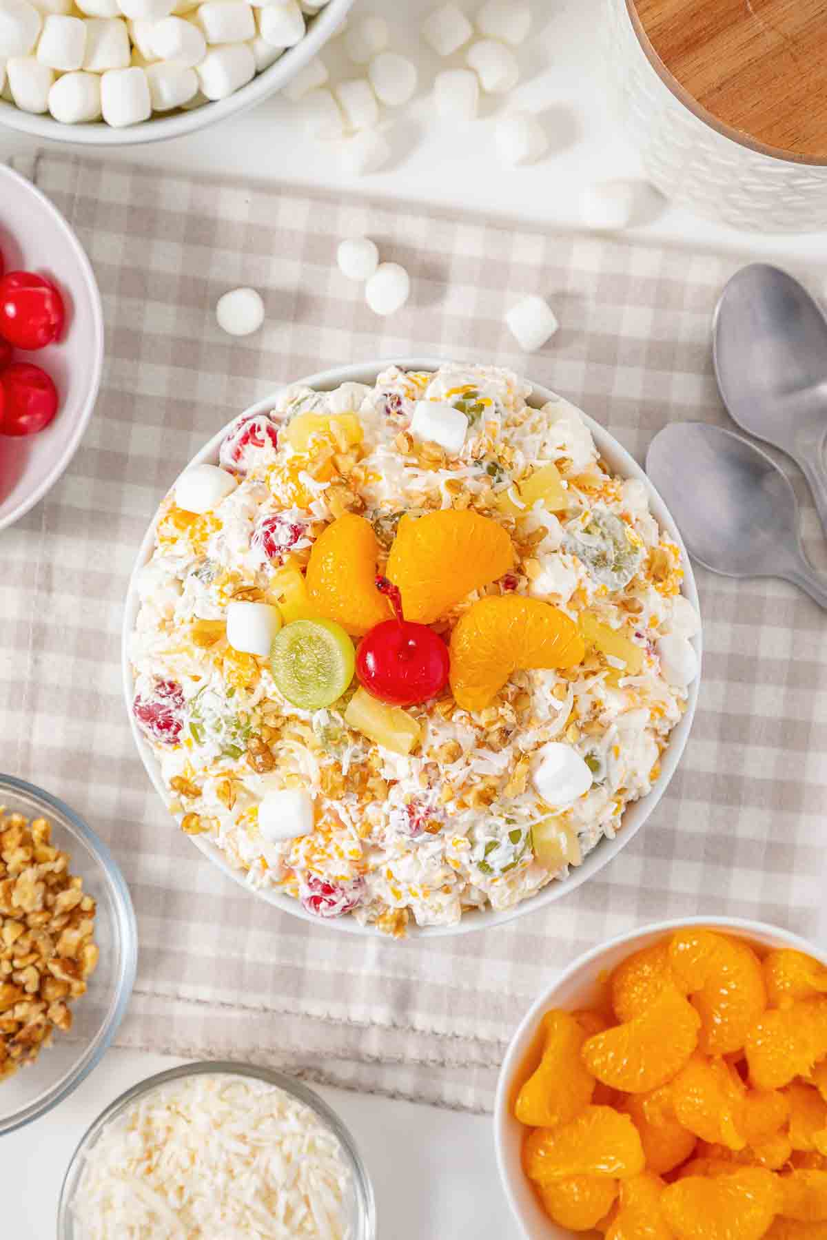 A bowl of ambrosia salad topped with fruit, cherries, and marshmallows sits surrounded by ingredients on a table, showcasing this classic fluff salad recipe.