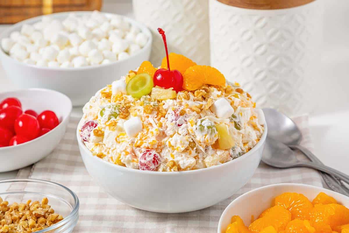 A bowl of Ambrosia Fluff Salad topped with a cherry, mandarin oranges, and coconut, surrounded by fresh ingredients.