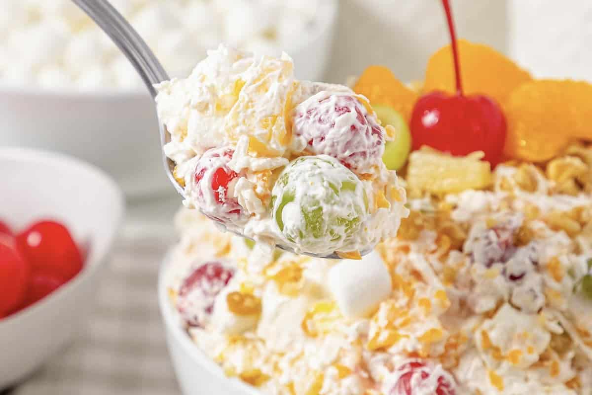 A spoonful of creamy Ambrosia fruit salad with grapes, cherries, and marshmallows rises above a bowl of the classic Fluff Salad.