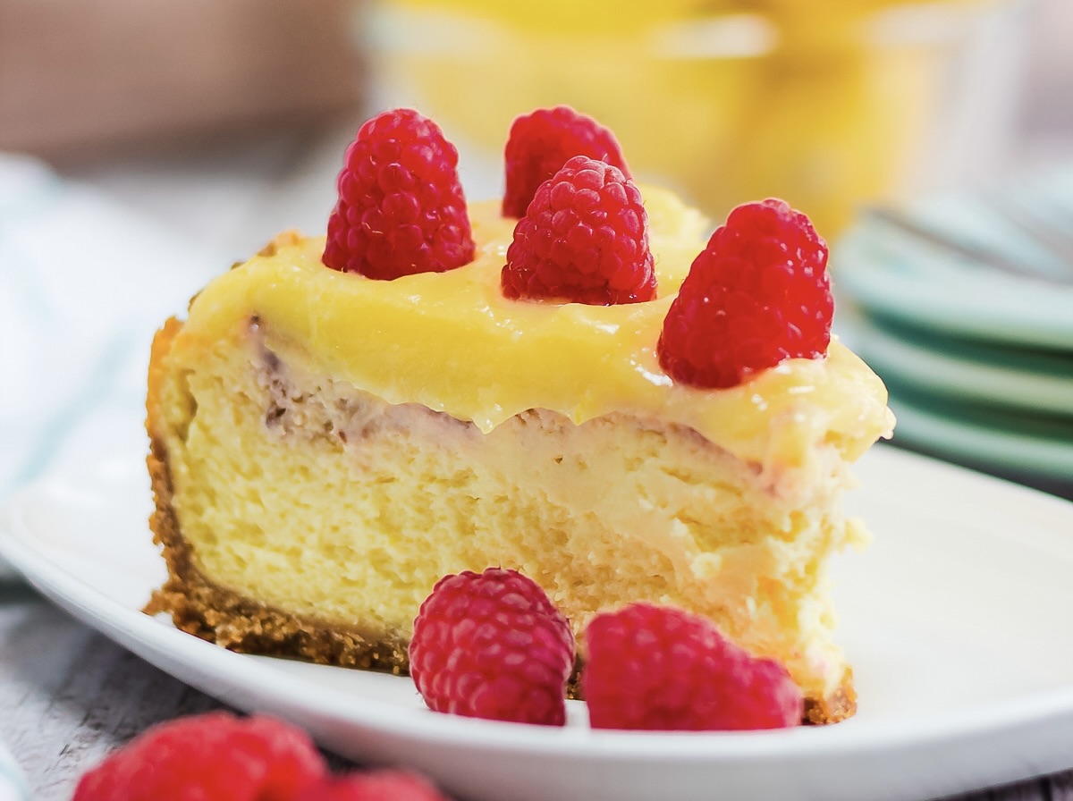 A slice of Raspberry Lemon Cheesecake topped with lemon curd and fresh raspberries on a white plate—try this irresistible cheesecake recipe for your next dessert craving.