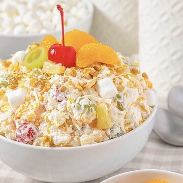 Ambrosia Fluff Salad topped with a cherry, orange slices, grapes, and marshmallows in a white bowl on a table.