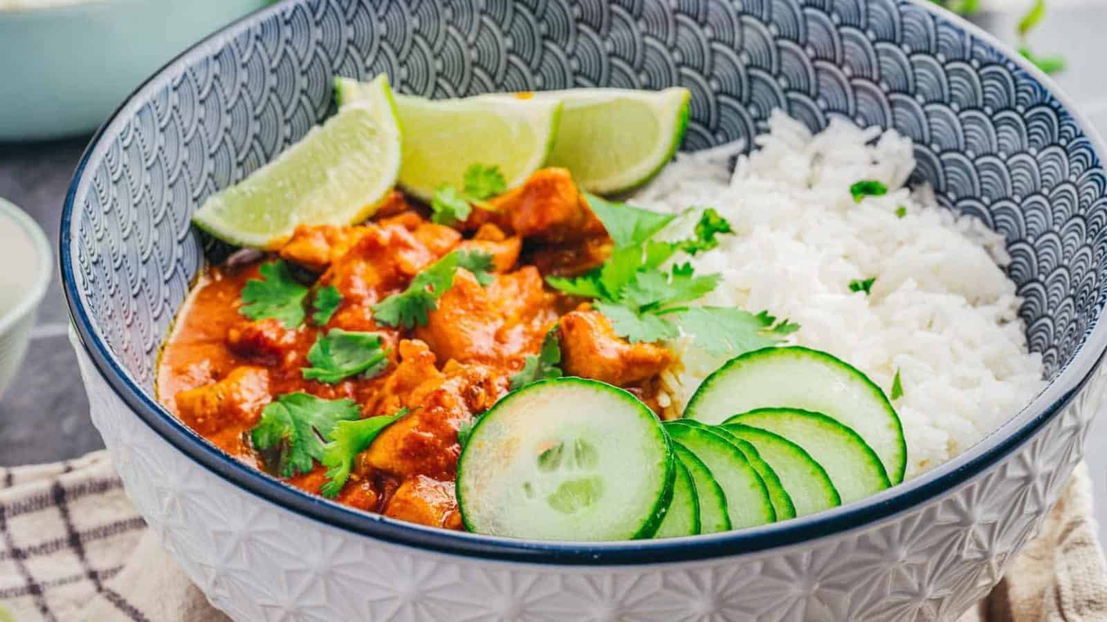 Bowl of curry with chicken, white rice, lime wedges, cucumber slices, and fresh cilantro on top.