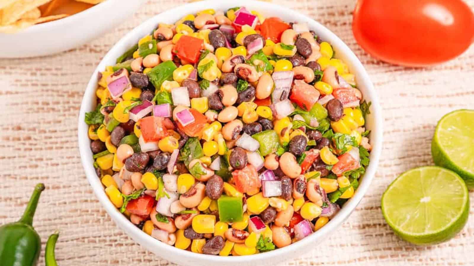 A bowl of colorful bean and corn salsa with chopped tomato, onion, cilantro, and lime on the side.