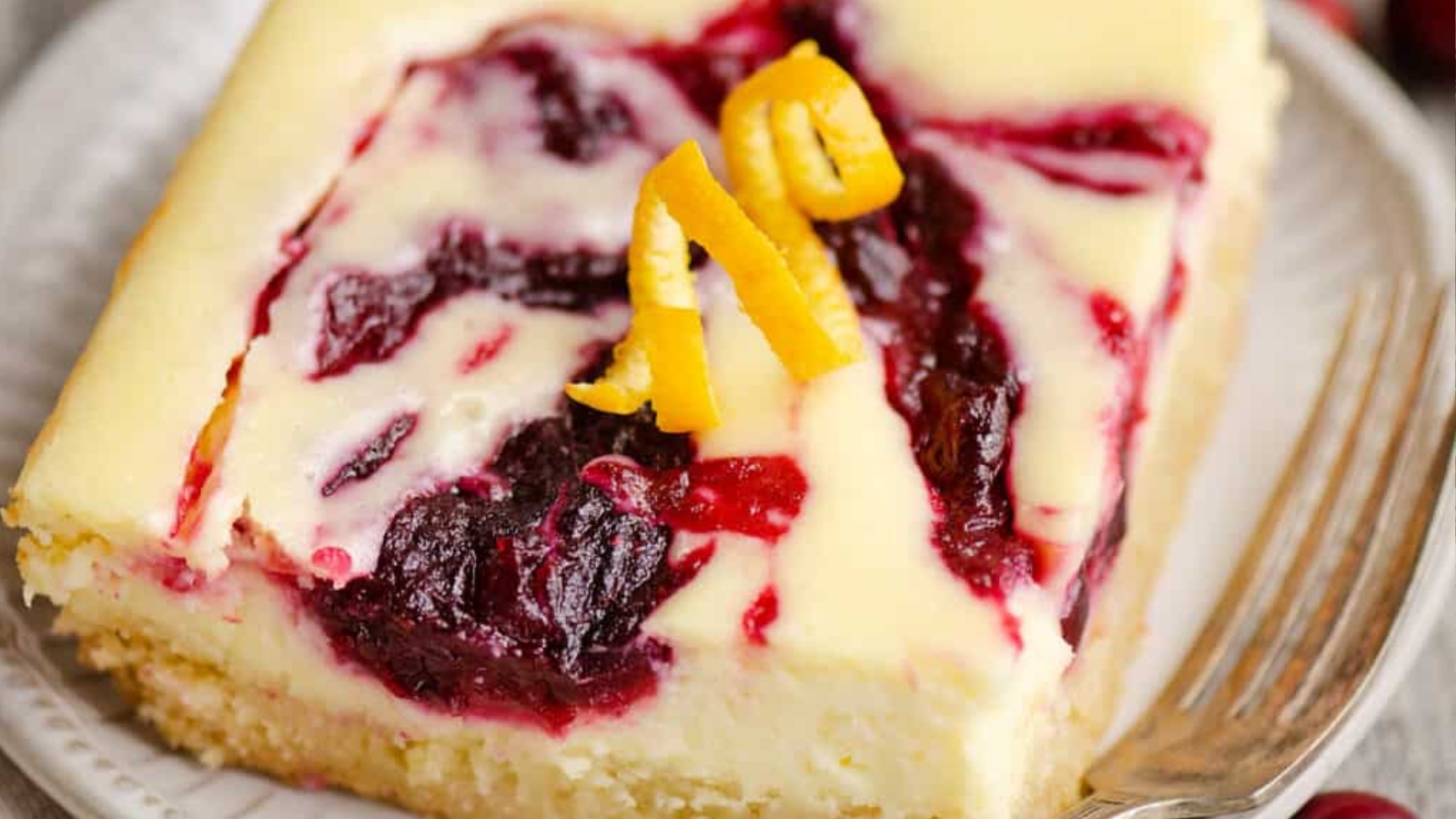 A cheesecake bar with berry swirl and orange zest topping on a white plate with a fork&mdash;perfect for budget Mother&rsquo;s Day desserts.