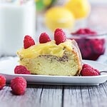 A slice of Raspberry Lemon Cheesecake with yellow topping and fresh raspberries on a white plate—perfect for trying a new recipe.