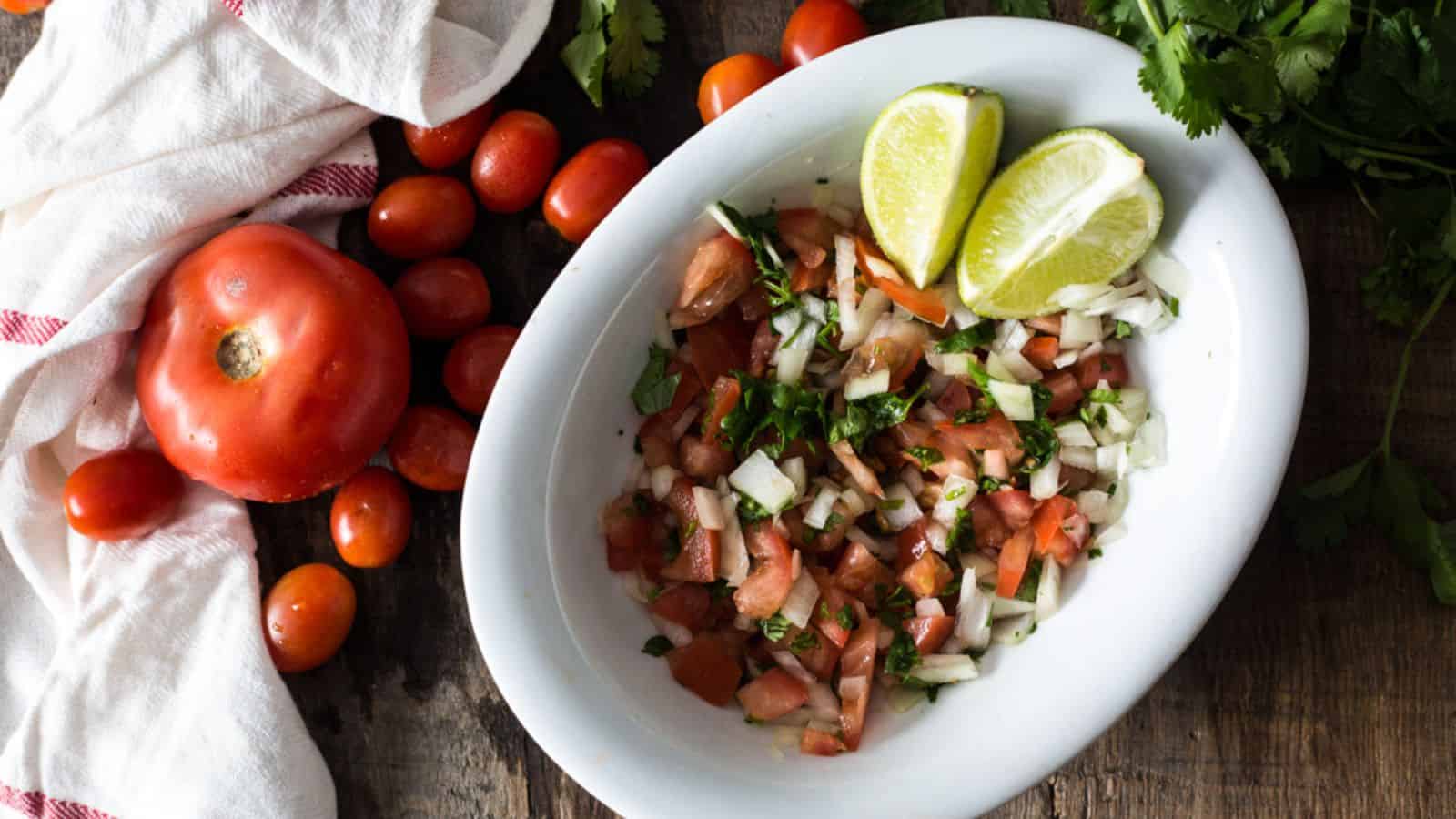 A bowl of pico de gallo with lime wedges, surrounded by fresh tomatoes and cilantro on a wooden table, brings vibrant flavor to your favorite Mexican recipes.
