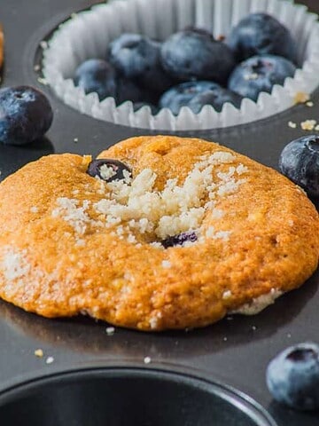 Blueberry muffins and fresh blueberries in a muffin tin, with crumbs sprinkled on top.