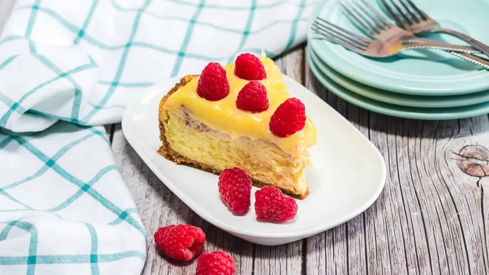 Slice of cheesecake topped with raspberries on a white plate, beside stacked plates, forks, and a checkered cloth.