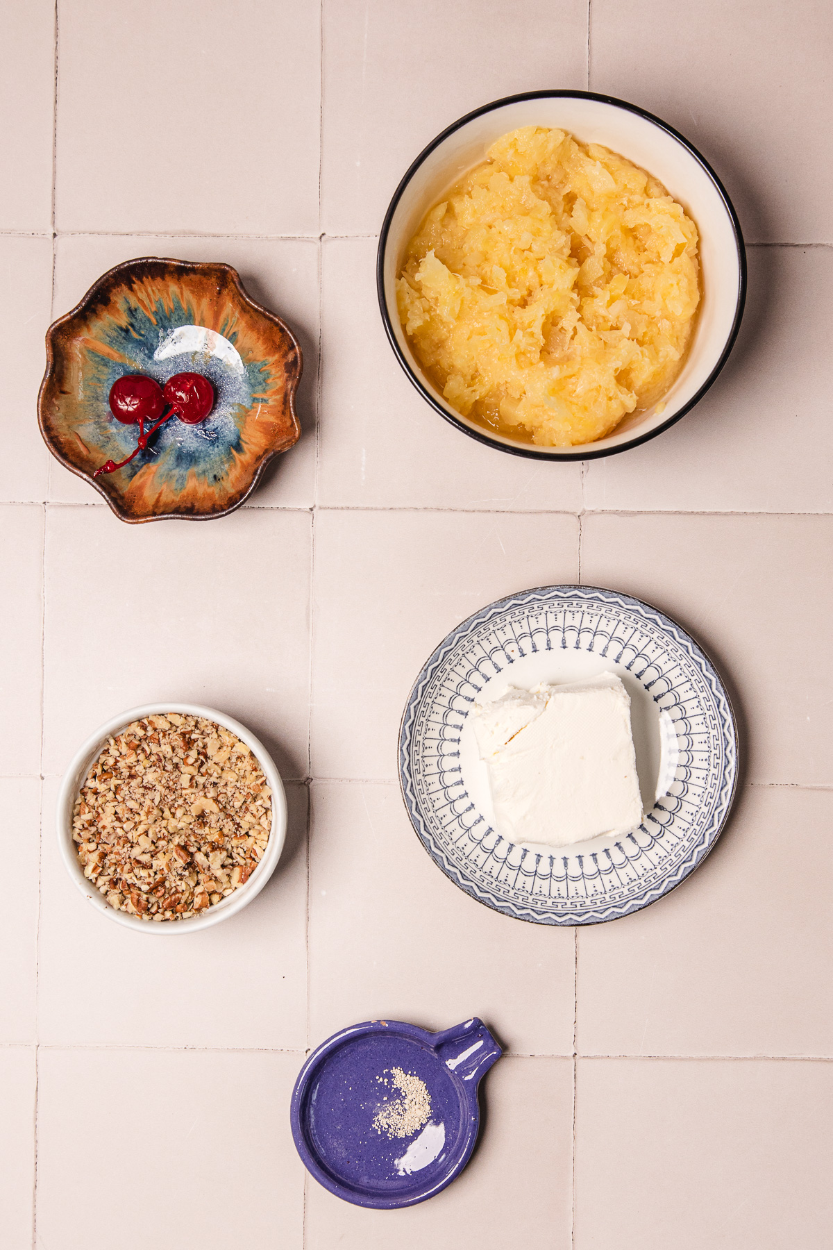 Five bowls with cherries, pineapple, cream cheese, chopped nuts, and a pinch of white spice on a tiled surface&mdash;perfect for assembling your favorite pineapple cheese ball recipe.