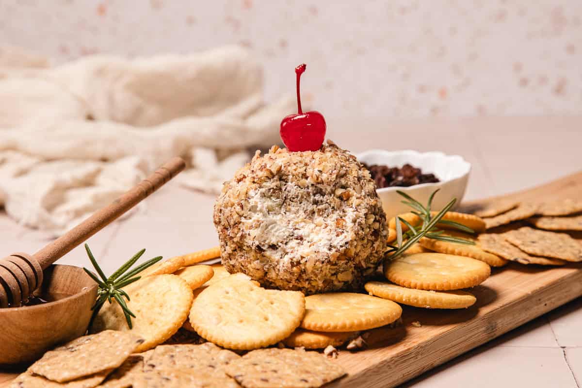 Cheese ball coated in nuts with a cherry on top, surrounded by crackers and rosemary on a wooden board&mdash;a delightful twist on the classic pineapple cheese ball recipe.