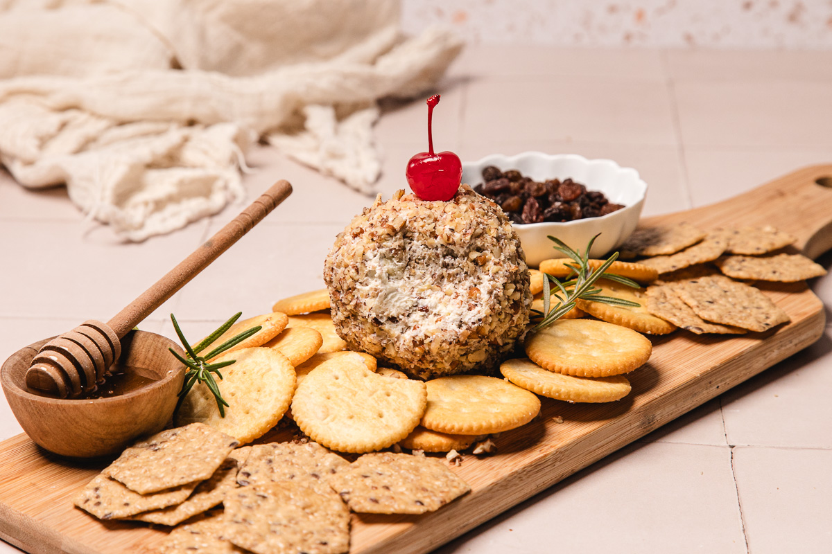 Cheese ball with a cherry on top, inspired by a pineapple cheese ball recipe, surrounded by assorted crackers, honey, and raisins on a wooden board.