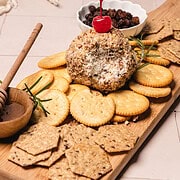 Cheese ball with crackers, honey dip, dried fruit, and a cherry on top, served on a wooden board—a delightful take inspired by the classic pineapple cheese ball recipe.