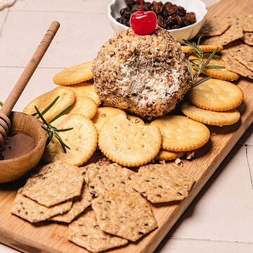 Cheese ball with crackers, honey dip, dried fruit, and a cherry on top, served on a wooden board&mdash;a delightful take inspired by the classic pineapple cheese ball recipe.
