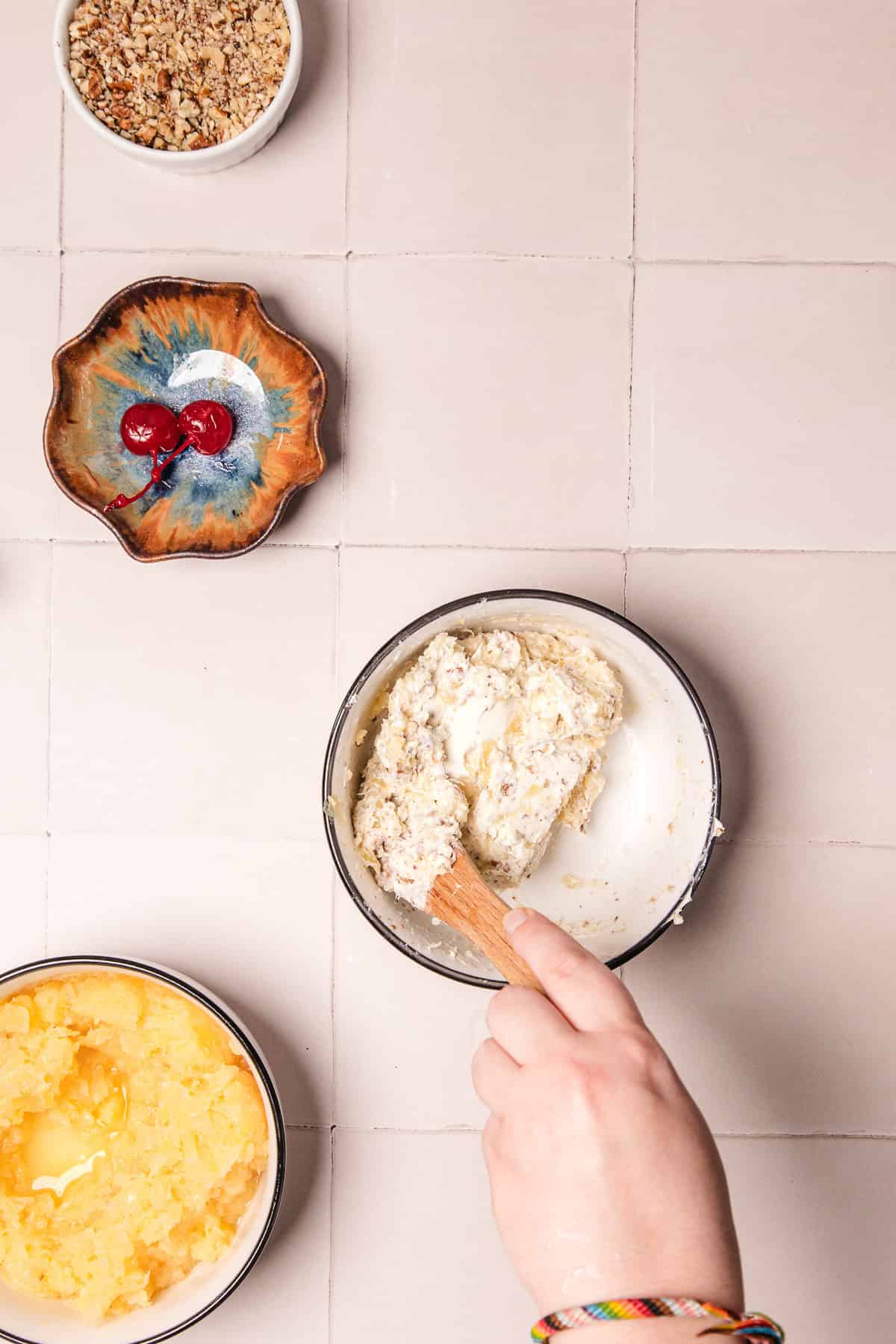 A hand mixes ingredients in a bowl near bowls of cherries, chopped nuts, and crushed pineapple on a tiled surface&mdash;perfect for preparing a classic pineapple cheese ball recipe.