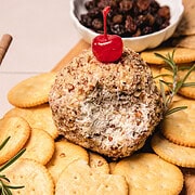 A pineapple cheese ball recipe coated in chopped nuts, topped with a cherry, and surrounded by crackers on a wooden board.