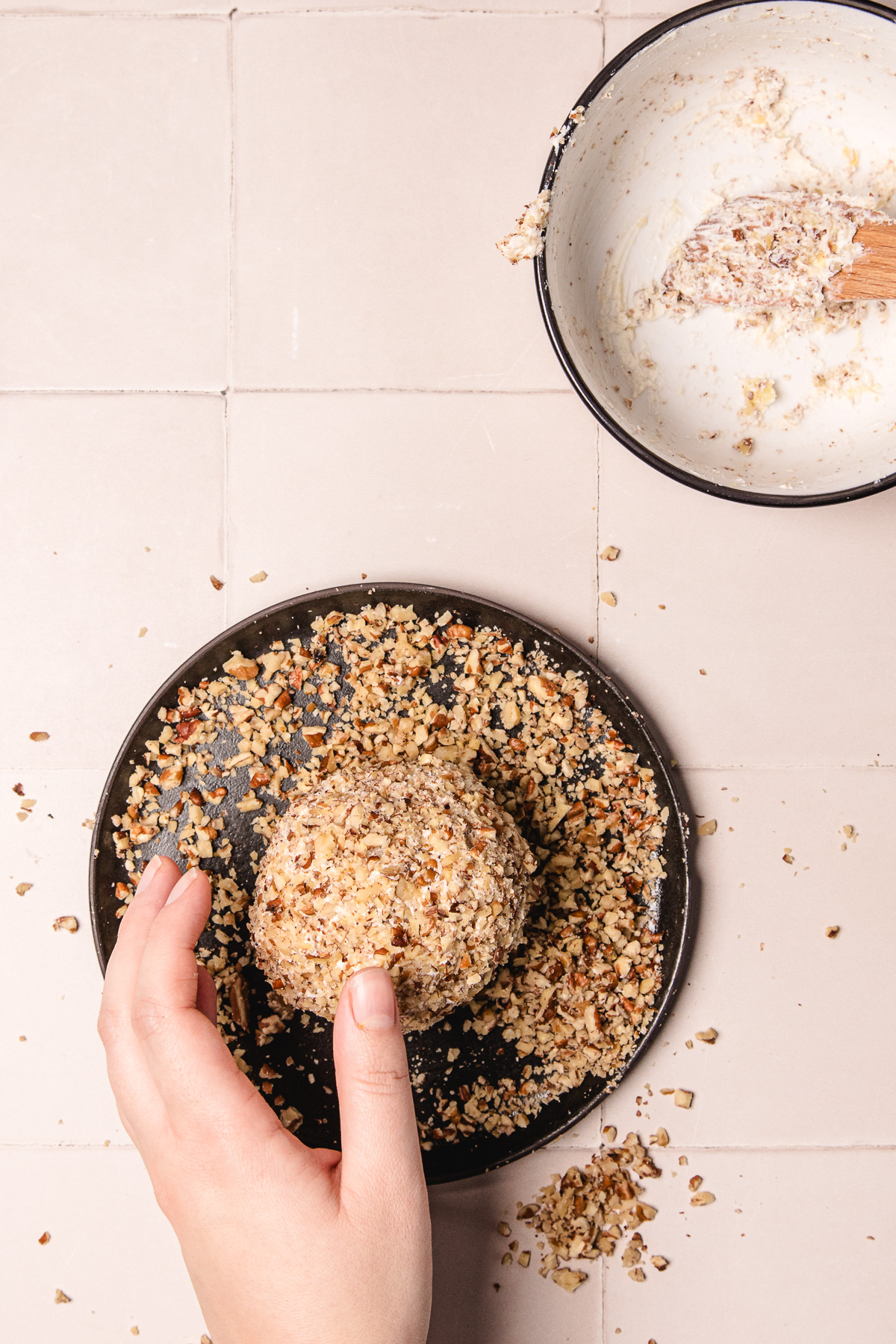 A hand rolls a pineapple cheese ball recipe in chopped nuts on a small plate beside a bowl with a wooden spoon.