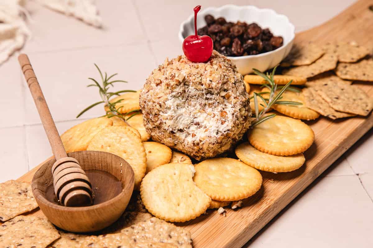 A pineapple cheese ball recipe coated in nuts, topped with a cherry, surrounded by crackers and a honey dipper on a wooden board.